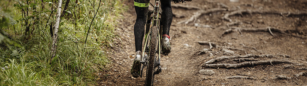 Ciclista com calça preta, pedalando em bicicleta em área rural com pouco de lama]