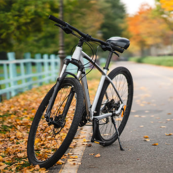 Bicicleta de cor predominante branca com detalhes em preto, apoiada em pezinho de encosto lateral, entre rua e canteiro com folhas, ao fundo cerca de madeira azul com árvores